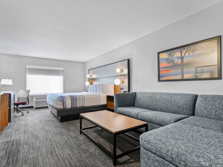 Een moderne hotelkamer met een bed, een grijze bank, een houten salontafel, een bureau met een computer en een groot raam met gordijnen.
