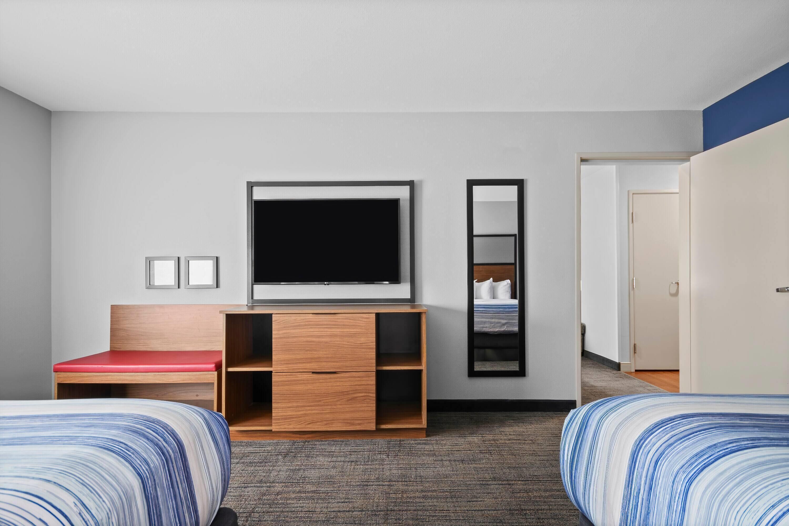 A hotel room with a bed, a TV, a desk with a red seat, a mirror, and a door.