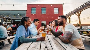 Un grupo de personas brindando en una mesa de madera al aire libre, con un edificio rojo al fondo.
