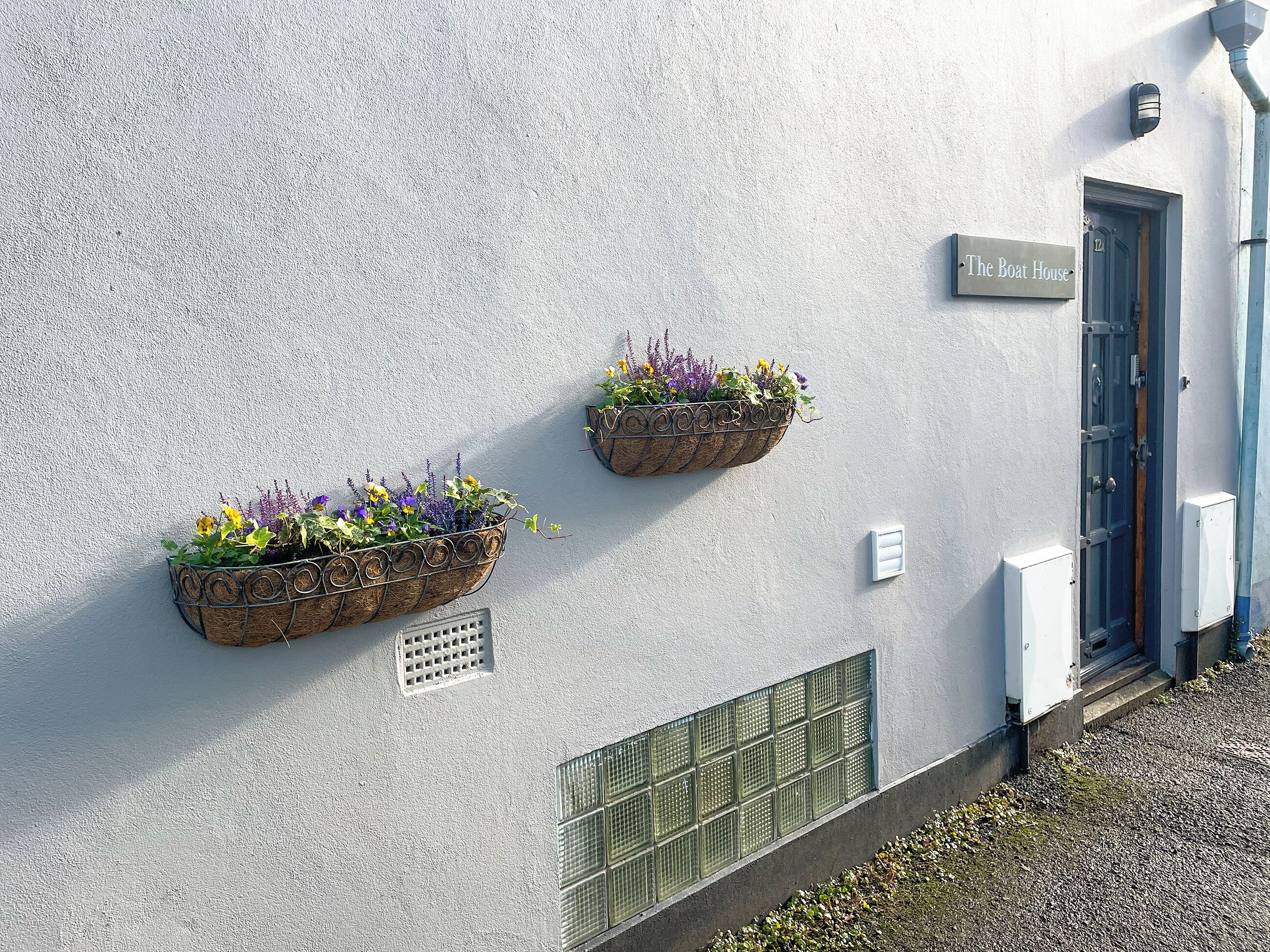Ein Gebäudefassaden mit einem Schild, das 'The Boat House' zeigt, zwei Blumenkästen, einer Tür und einigen Stromverteilern.