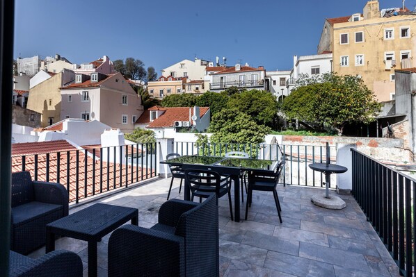 Terraço com mobiliário de exterior, mesa e cadeiras, com vista para um horizonte urbano repleto de edifícios e árvores.