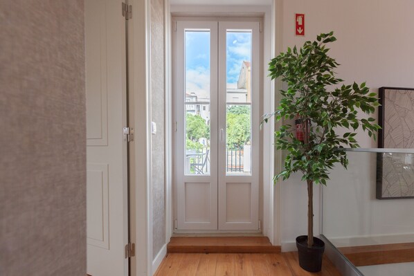 Un pasillo con suelo de madera, una planta en maceta y una puerta que da a un balcón con vistas a edificios y árboles.