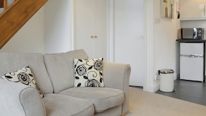 Una sala de estar con un sofá gris, cojines estampados, una escalera de madera, una puerta blanca, una cocina con un microondas y un cubo de basura.
