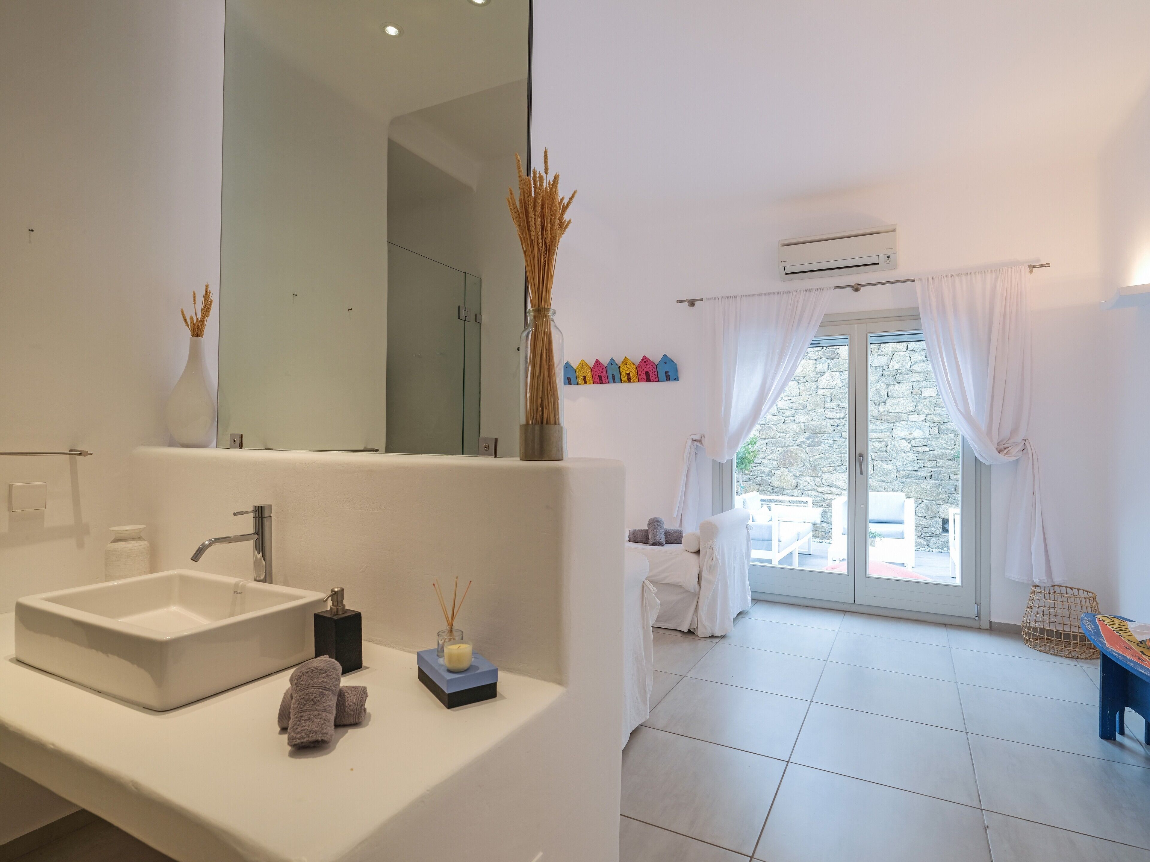 Un baño moderno con un espejo grande, un lavabo blanco y una zona de ducha. Hay una ventana con cortinas y vistas al exterior.
