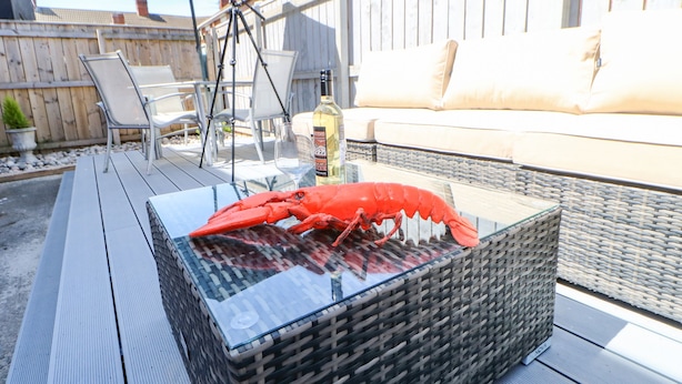 A wicker table with a glass top, a red lobster, outdoor furniture, and a wooden fence in the background. - Lobster Pot Retreat (Newbiggin-by-the-Sea)