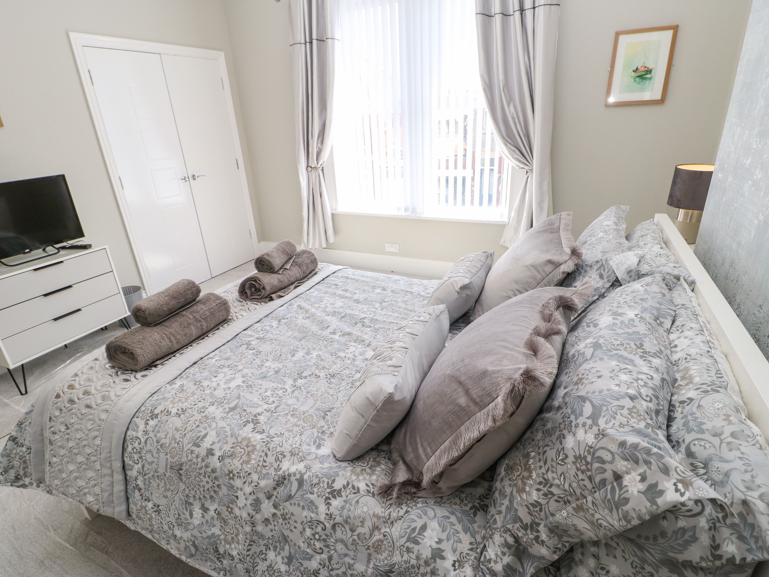 A neatly made bed with patterned bedding, a television on a white dresser, and a window with curtains.