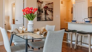 Un coin repas avec une table en verre, des chaises grises, un vase de tulipes rouges et un tableau représentant une scène de rue.