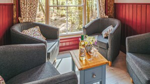 Een kamer met twee grijze fauteuils, een kleine houten tafel met een fles en glazen, en een raam met uitzicht op een bos.
