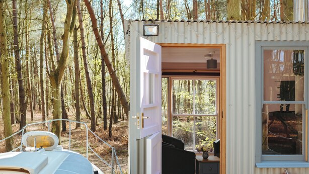 Een klein huisje met een deur naar een interieur met een bank, een tafeltje en een raam.