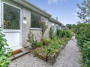 A row of single-story buildings with large windows, a gravel pathway, and various plants and shrubs.