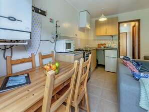 A compact kitchen with a microwave, stove, and refrigerator. There is a dining table with chairs and a bowl of fruit. A sofa is visible in the background. - 20 Blackbird Cottage-UK38935 (Lands End, near Porthcurno)