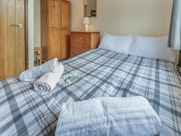 A neatly made bed with a plaid bedspread, a wooden wardrobe, a dresser, and a mirror on the wall.