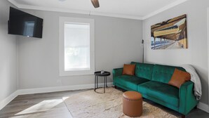 Un soggiorno con un divano verde, un poggiapiedi marrone, una ventola a soffitto, una TV a parete e un'ampia finestra con tende.