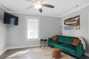 Ein Wohnzimmer mit einem grünen Sofa, einem braunen Hocker, einem Deckenventilator, einem Wand-Fernseher und einem großen Fenster mit Jalousien.