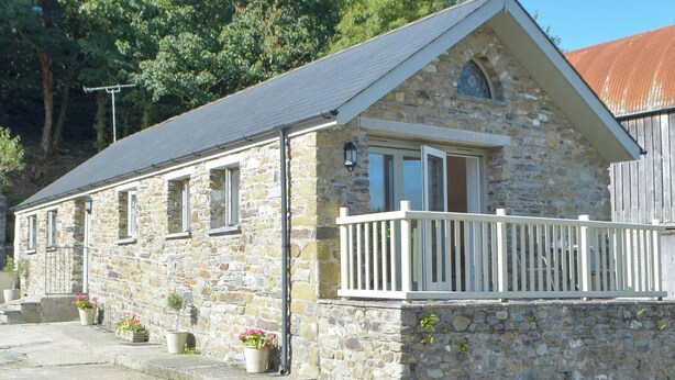 A stone building with a gabled roof, a balcony with white railings, and potted plants on the ground floor. - Troed Y Rhiw Aberpoth (Aberporth)