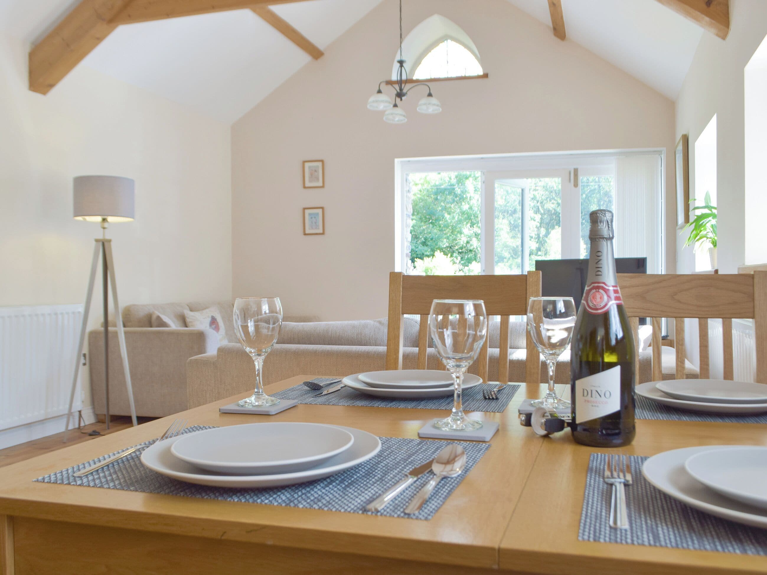 A dining area with a table set for four, a bottle of DINO Prosecco, and a view of a cozy living room with a sofa and a lamp.