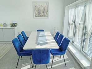 Un comedor moderno con una mesa blanca y sillas azules, un armario blanco con utensilios de cocina, y un ventanal con cortinas traslúcidas.
