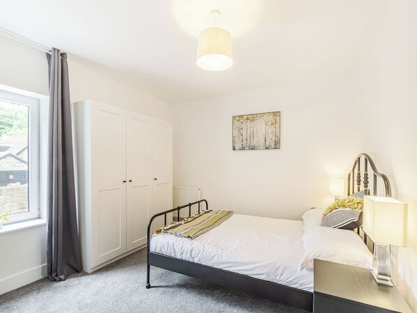 Ein Schlafzimmer mit Bett, Kleiderschrank, Heizkörper, einem Gemälde und einem Fenster mit Vorhängen.