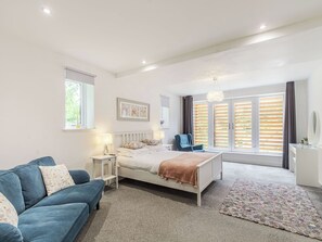 A bedroom with a bed, a blue sofa, a bedside table, a chair, a window with blinds, and a hanging light fixture.