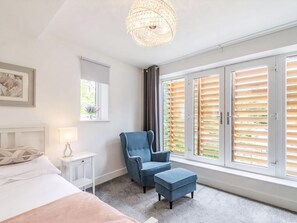 Une chambre à coucher comprenant un lit, une chaise, un pouf, une table de chevet, une lampe, une fenêtre avec des stores et un lustre.