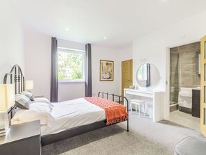 A hotel room with a bed, bedside table, lamp, window, desk, chair, mirror, and a shower area.