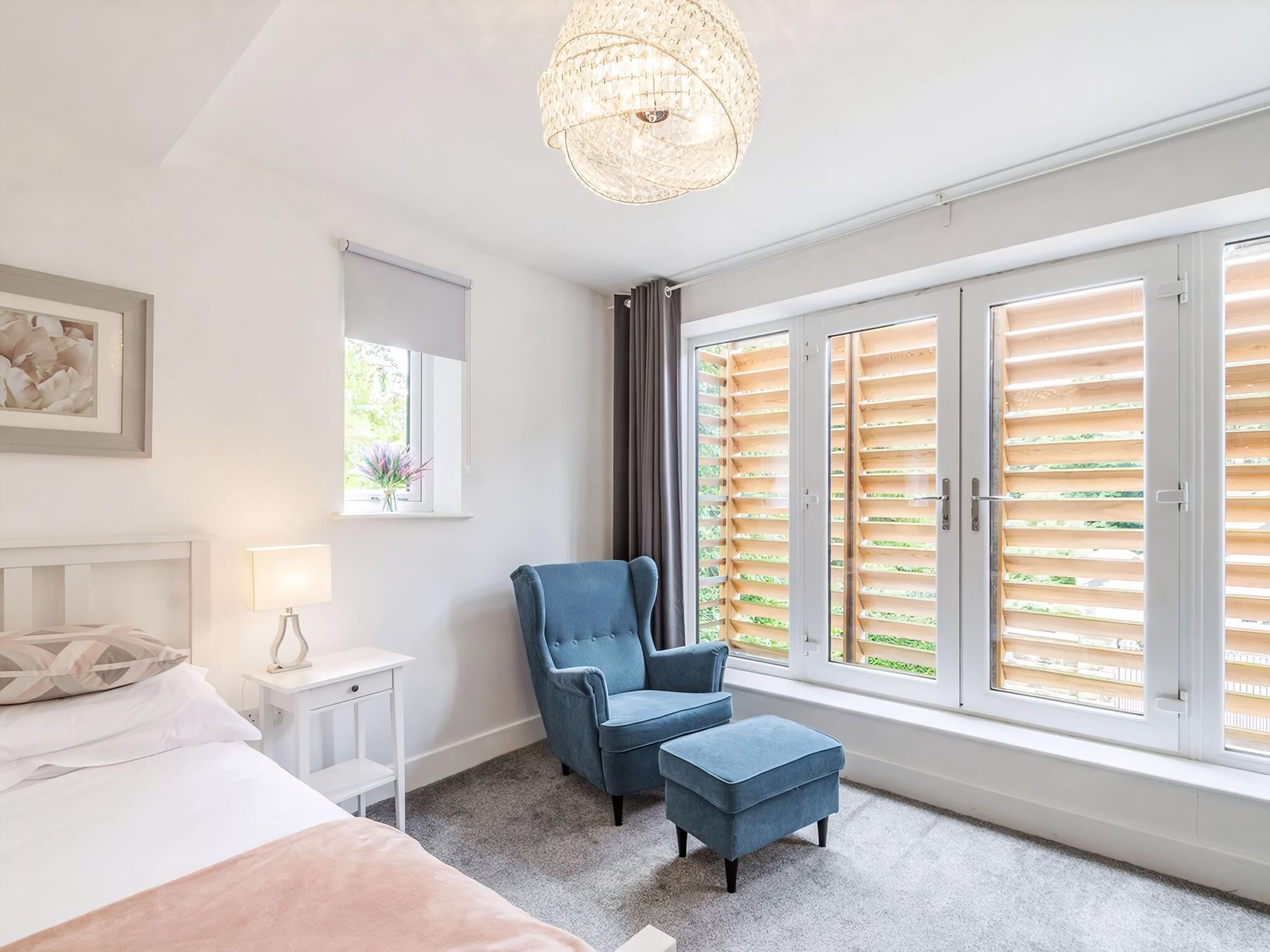 A bedroom with a bed, a chair, an ottoman, a nightstand, a lamp, a window with blinds, and a chandelier.