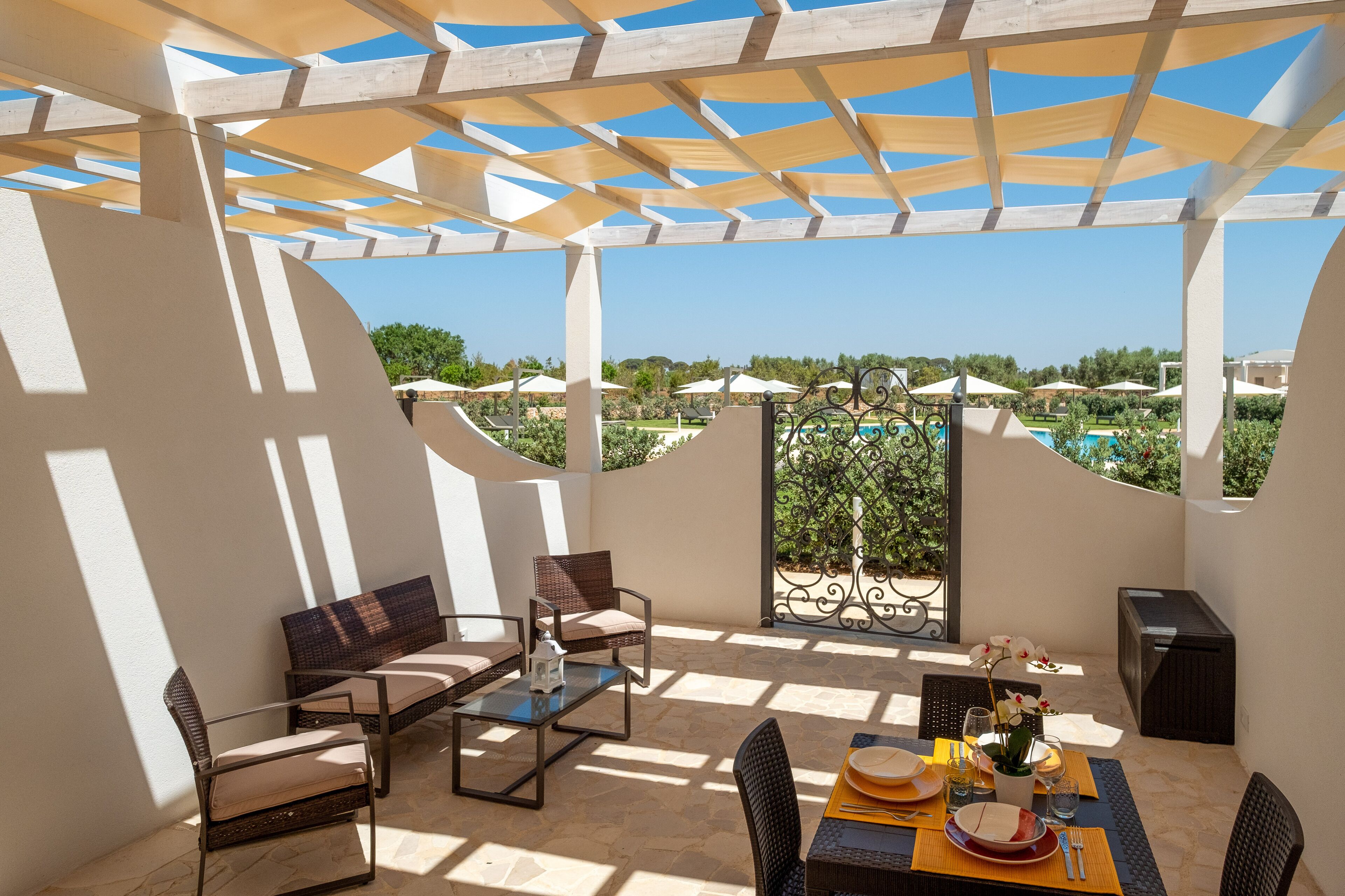 An outdoor patio with a table set for dining, surrounded by chairs, and a view of a pool and greenery.