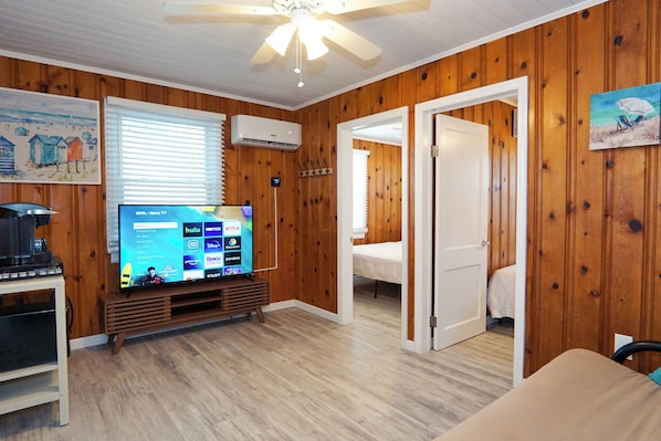 A room with wooden walls, a ceiling fan, a TV on a wooden stand, and a bed in the adjacent room.