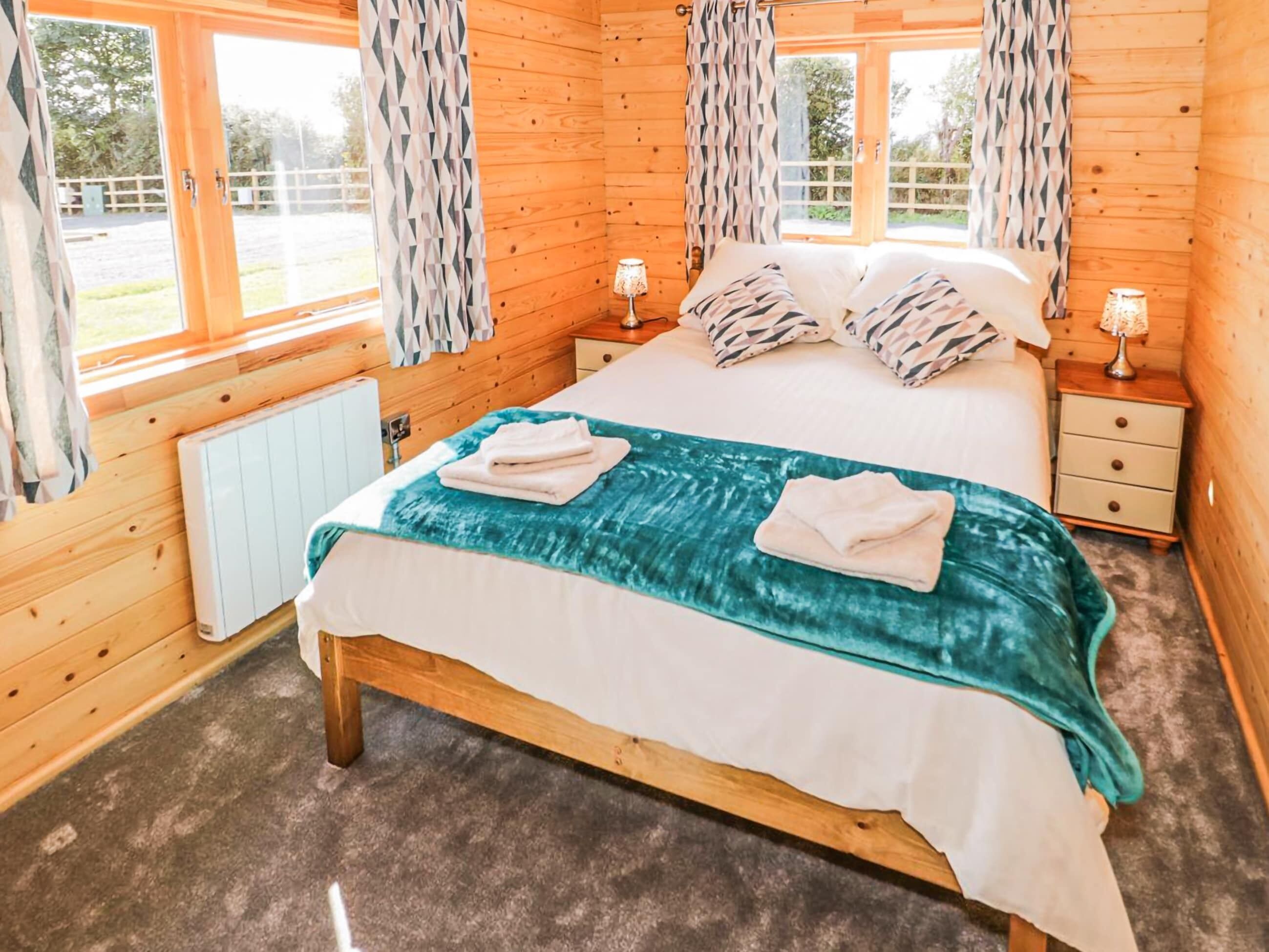 Un dormitorio de cabaña de madera con una cama, dos mesitas de noche, un radiador y cortinas.