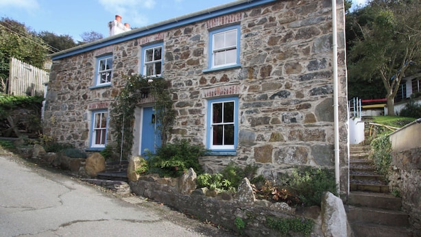 A stone building with a blue door and windows, surrounded by vegetation and a paved road.