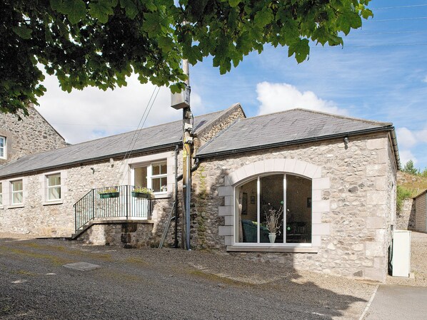 A stone building with a large arched window, a staircase leading to a balcony, and a gravel driveway. - 3 bedroom accommodation in Wooler (Wooler)