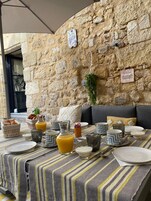 Una mesa para el desayuno con un fondo de pared de piedra, mantel a rayas y diversos productos para el desayuno.