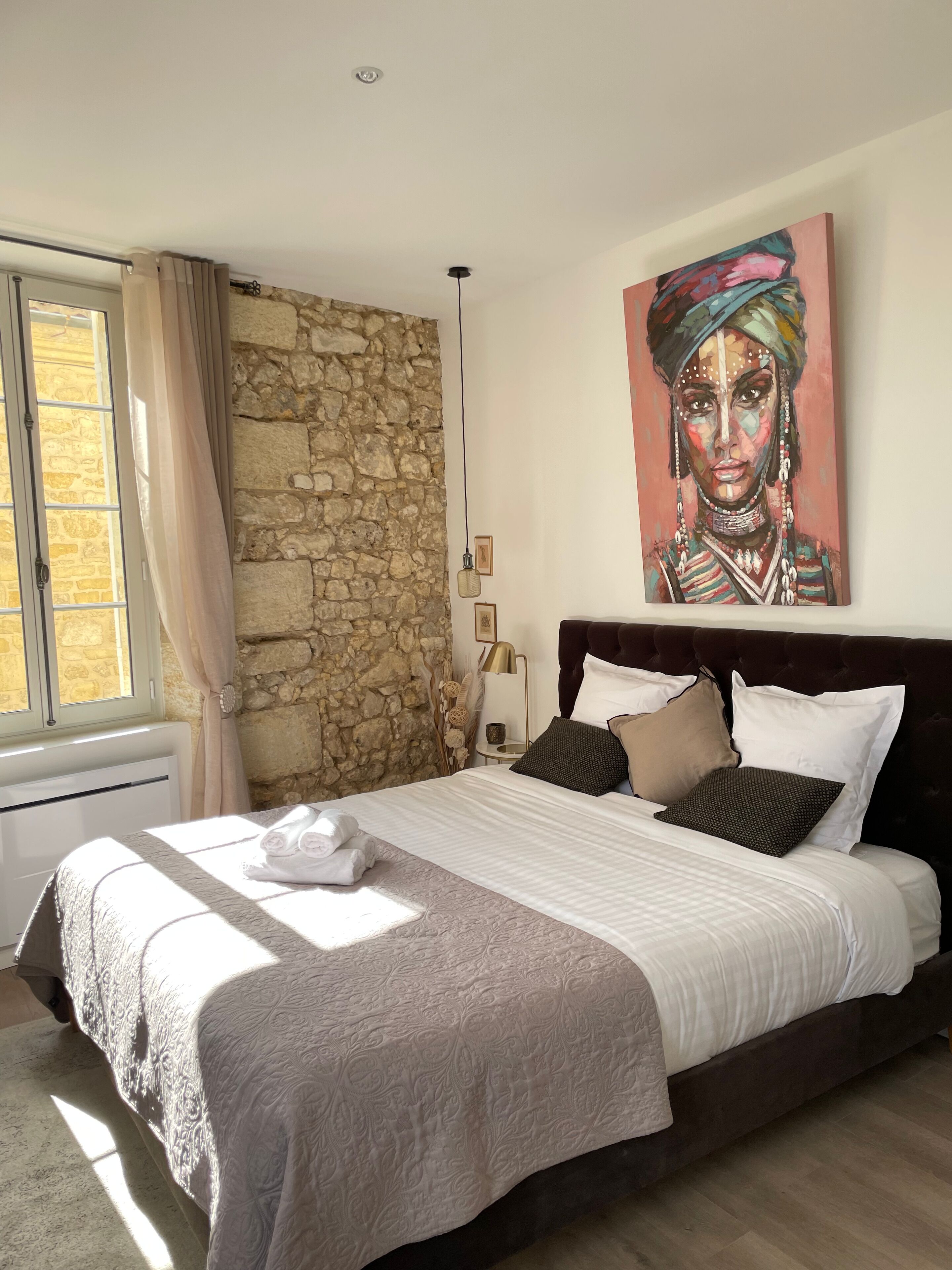 Un dormitorio con pared de piedra, una cama grande con muchos almohadones, un cuadro en la pared y una ventana con cortinas.