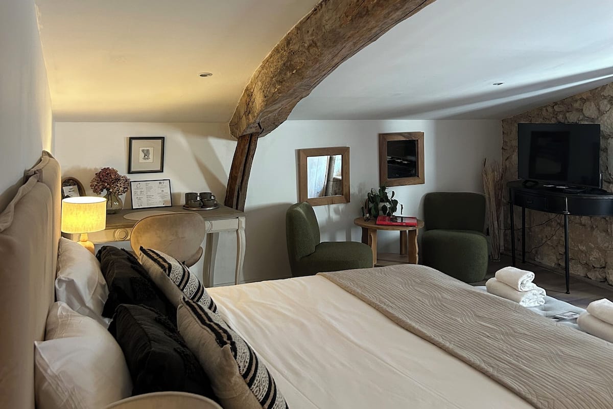 A bedroom with a large bed, a desk, a chair, a television, and a stone wall.
