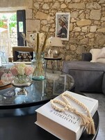 Una sala moderna con pared de piedra, una mesa de centro de cristal, un libro, un collar de cuentas y un jarrón con plantas secas.