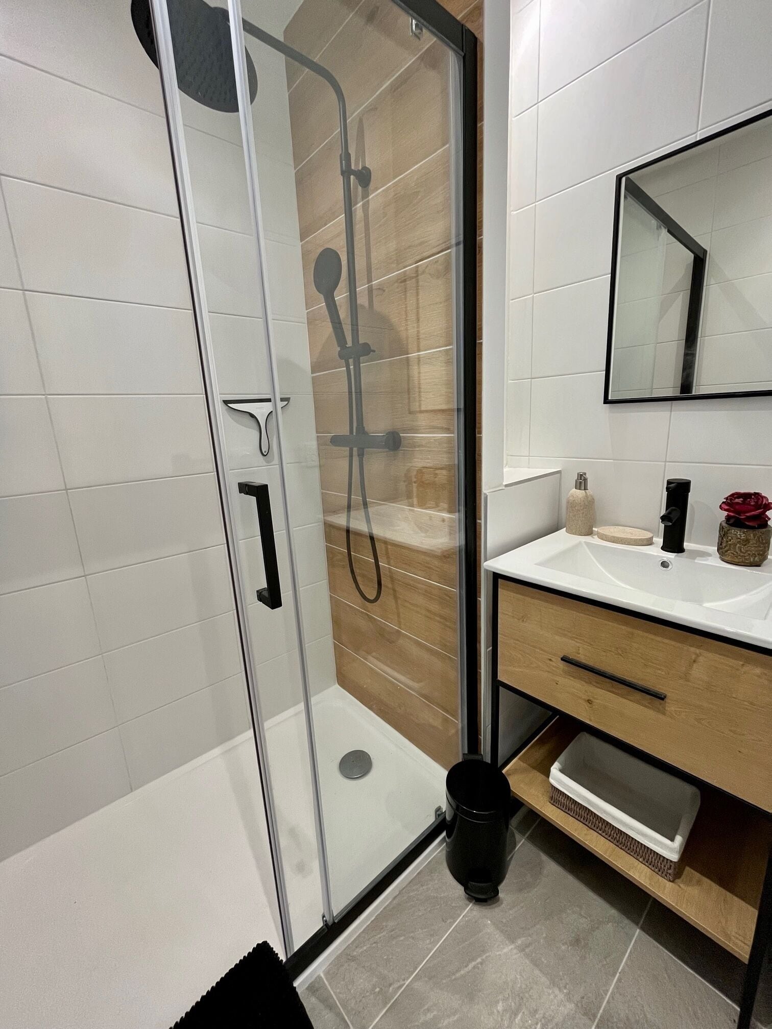 Ein modernes Badezimmer mit Duschkabine aus Glas, weißen Fliesen, einem Holzwaschtisch und einem Spiegel.