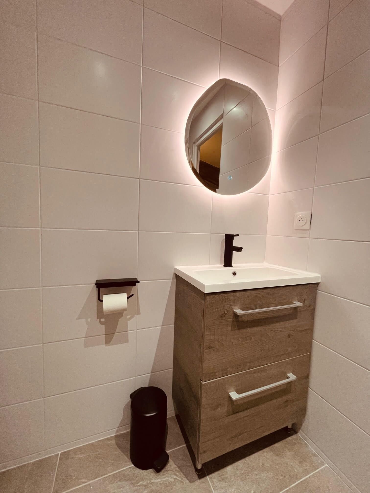 Un baño con tocador de madera, un espejo redondo con iluminación, un dispensador de papel higiénico y un cubo de basura.