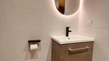 Un baño con tocador de madera, un espejo redondo con iluminación, un dispensador de papel higiénico y un cubo de basura.