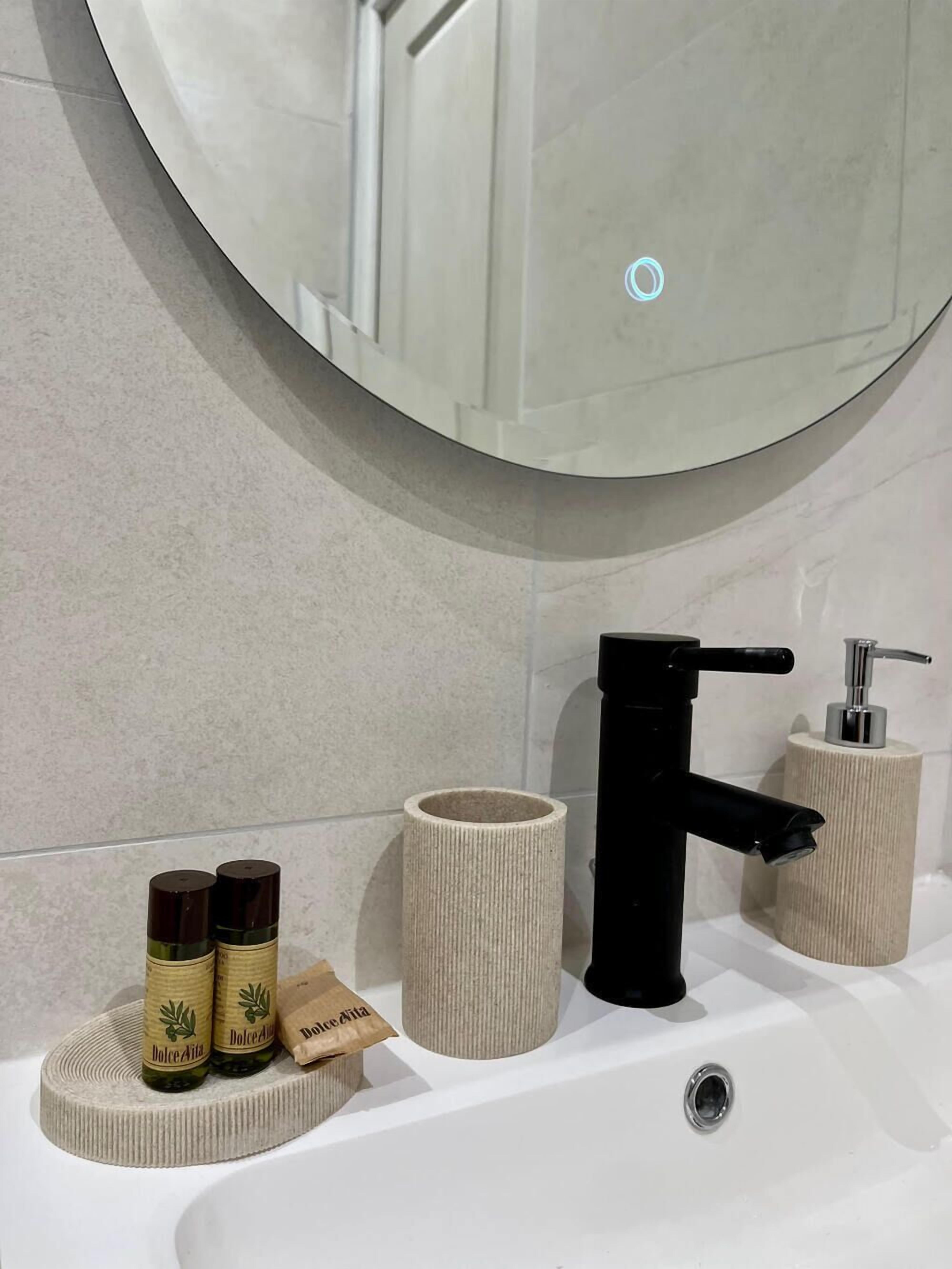 Un baño con un espejo redondo, un grifo negro y dos botellas de aceite de oliva Dolce Vita sobre un plato para jabón beige texturizado.