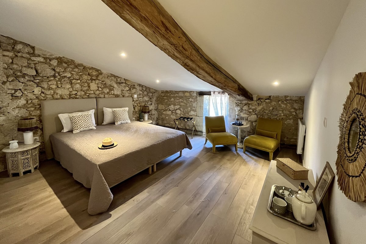 A bedroom with a stone wall, a large bed, a wooden ceiling beam, a small table with a lamp, a chair, and a tray with tea set.