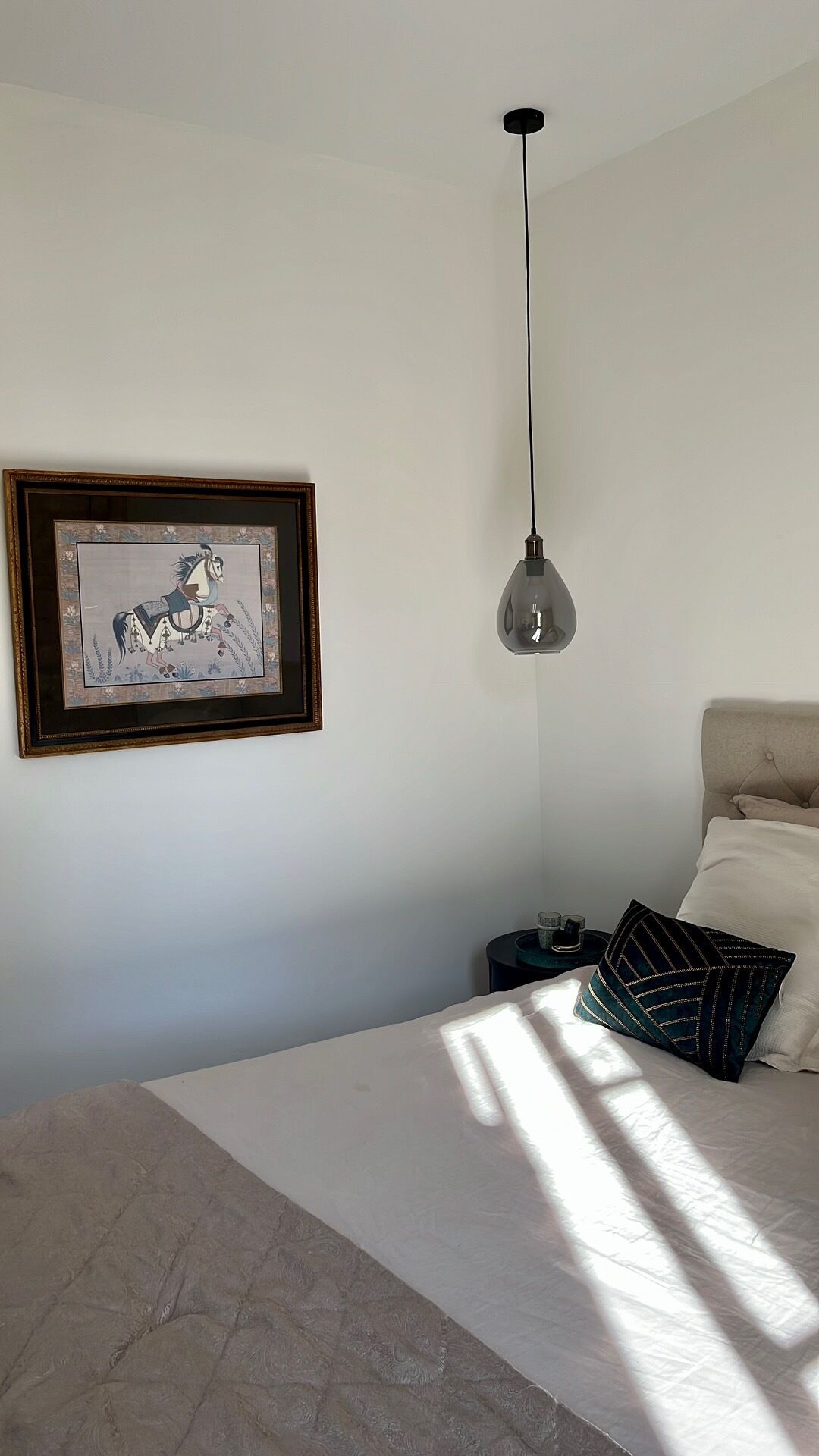 A bedroom with a bed, a hanging light fixture, a framed picture on the wall, and a bedside table with a lamp.
