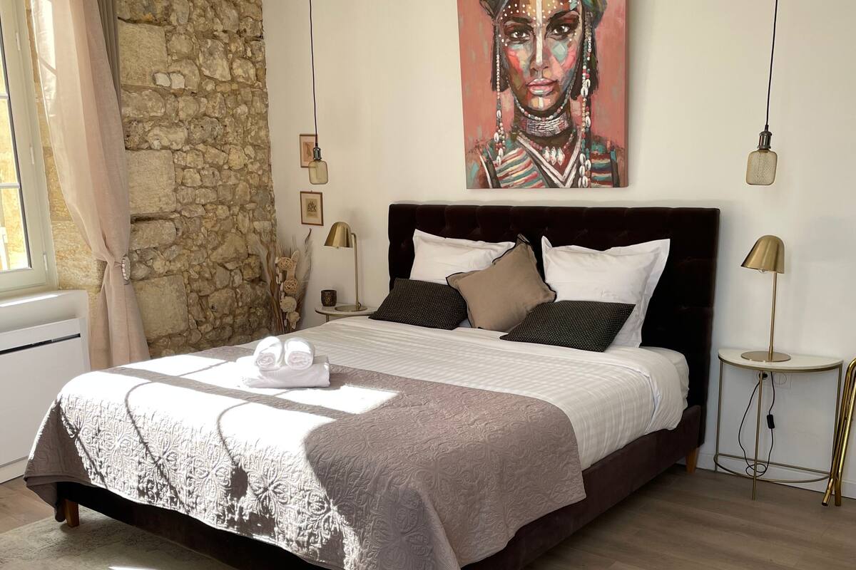A modern bedroom with a stone wall, a large bed with multiple pillows, a painting above the bed, and a small side table with a lamp.