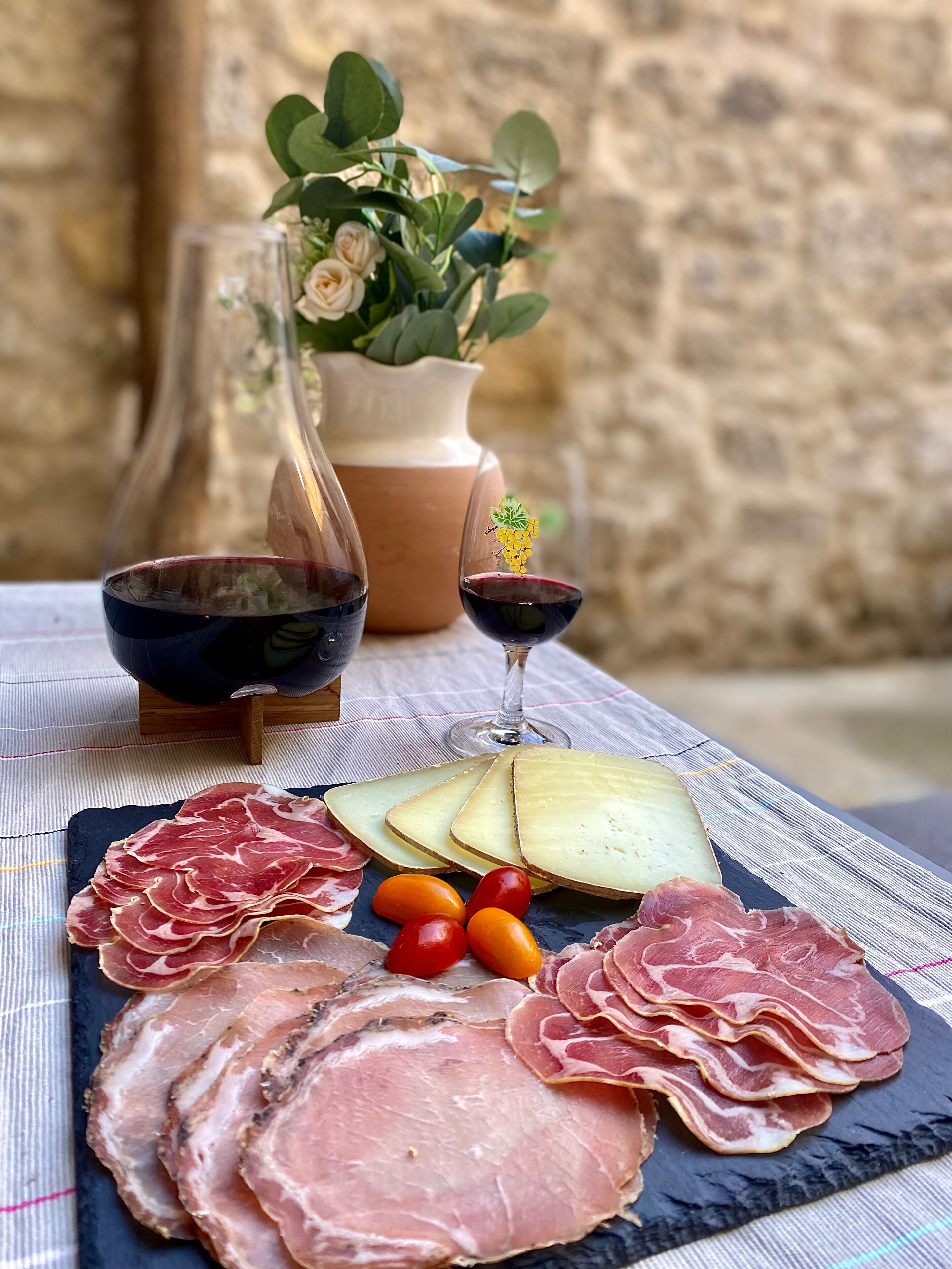 A charcuterie board with various meats, cheeses, and tomatoes on a slate board, accompanied by wine glasses and a vase of flowers.
