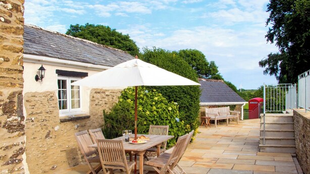 A stone building with a patio area featuring a wooden table and chairs, a white umbrella, and a red mailbox.