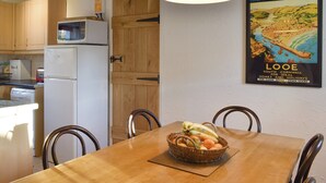 A dining area with a wooden table, chairs, and a basket of fruit. A white refrigerator and a microwave are visible in the background.