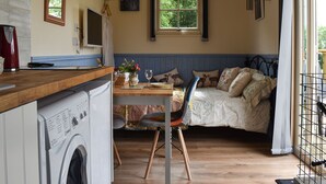 A compact room with a washing machine, a dining table with chairs, a bed with patterned pillows, a window with a view of greenery, and a small kitchen area. - 1 bedroom accommodation in Herstmonceux, near Hailsham (Herstmonceux, near Hailsham)