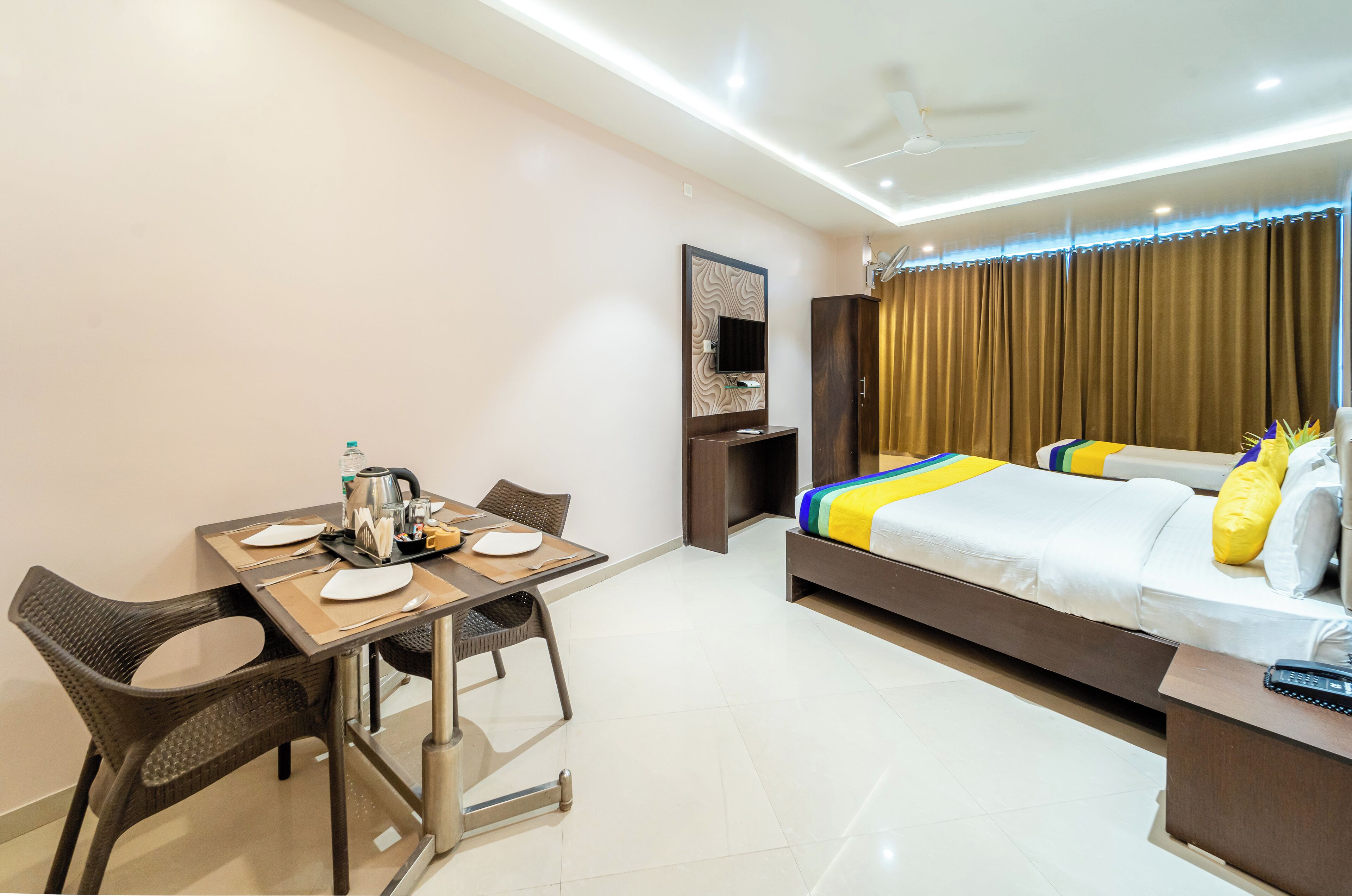 A hotel room with a bed, a desk with a chair, a table with a chair, a television, and a window with curtains.