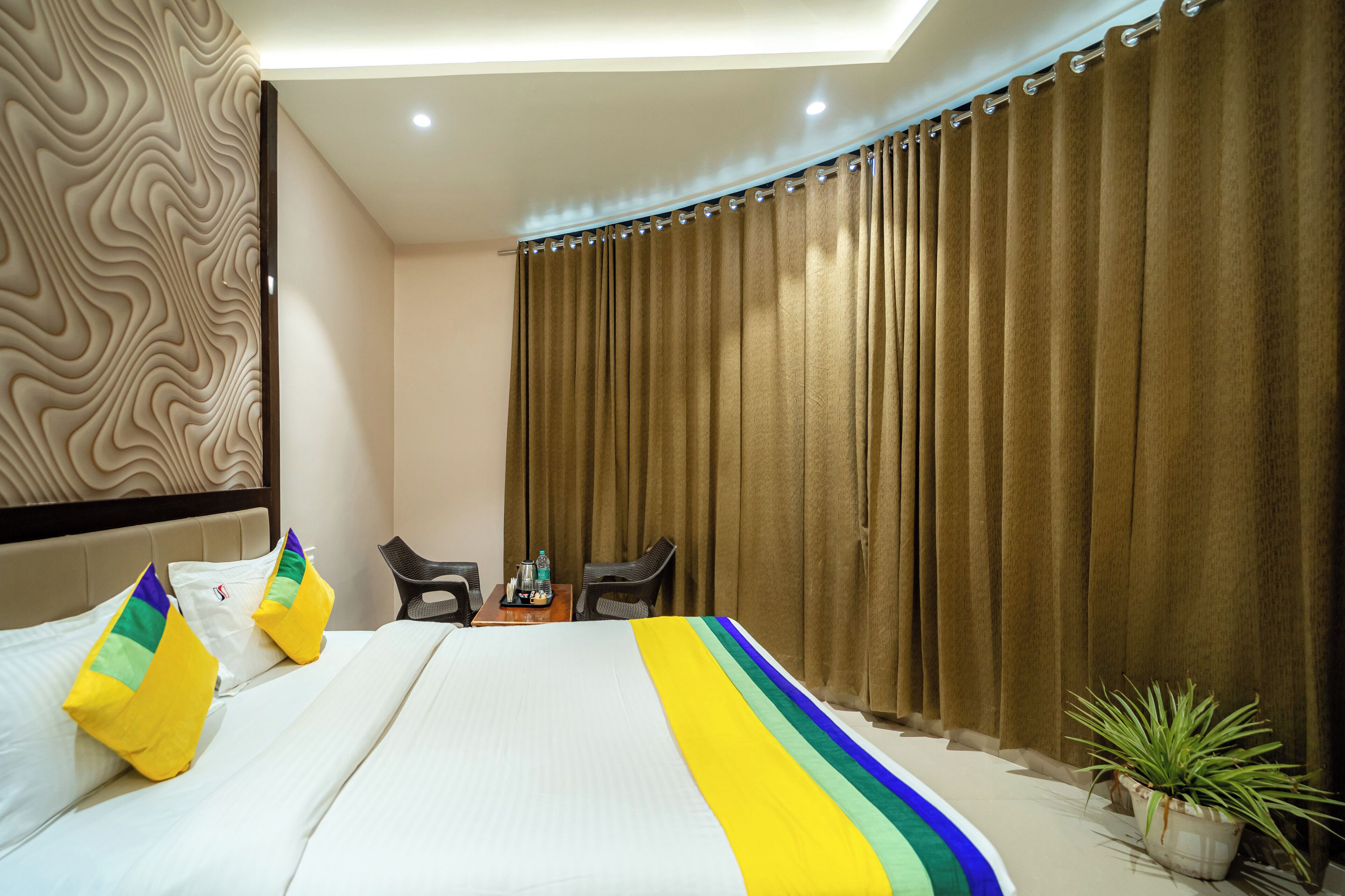 A hotel room with a bed, two chairs, a small table with a bottle and a glass, and a potted plant.