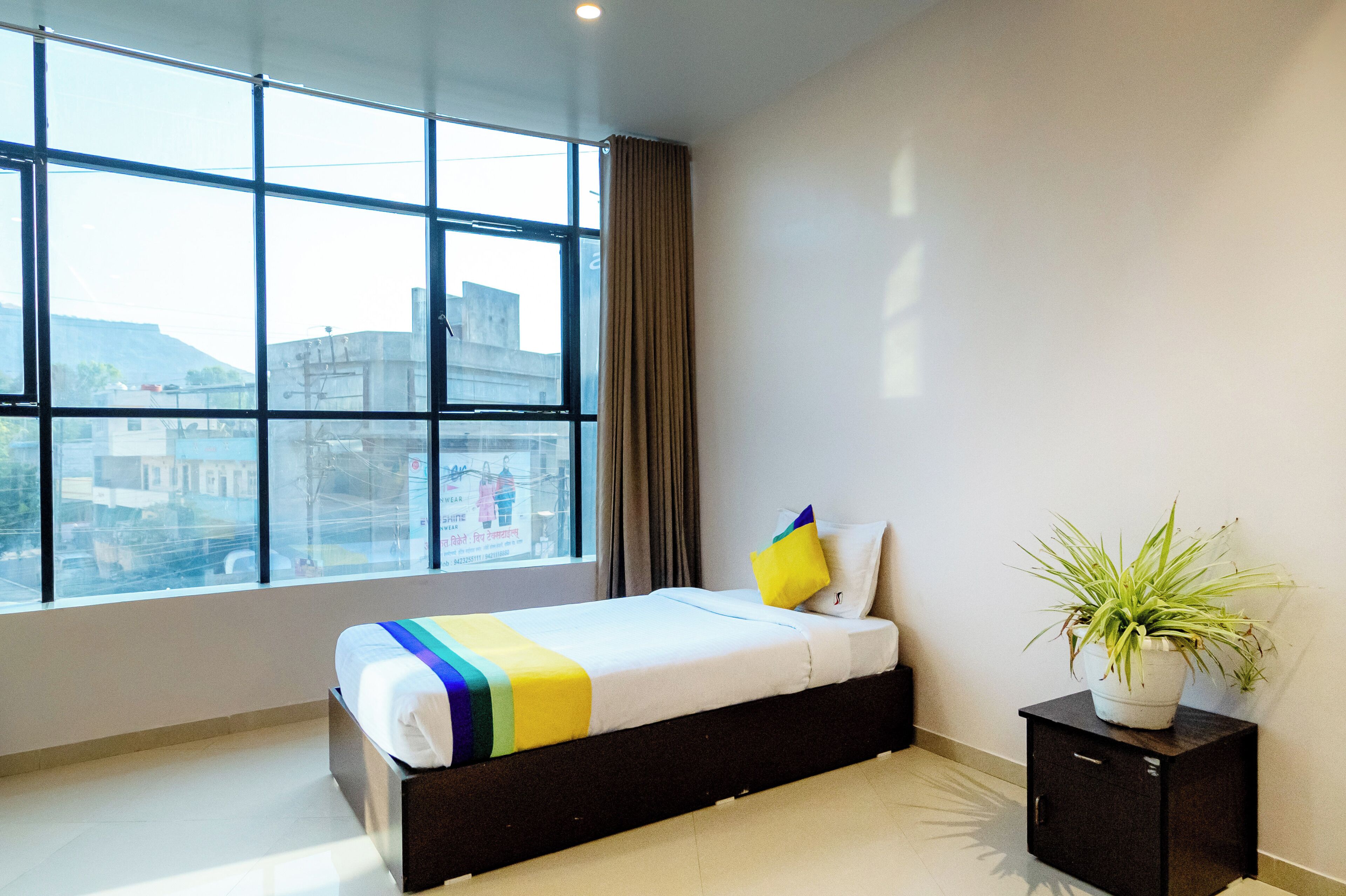 A hotel room with a large window, a bed with a colorful blanket, a potted plant, and a small cabinet.