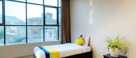 A hotel room with a large window, a bed with a colorful blanket, a potted plant, and a small cabinet.